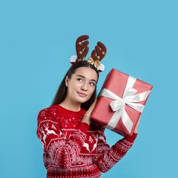 Young woman in Christmas sweater and reindeer headband holding gift on light blue background. Space for text