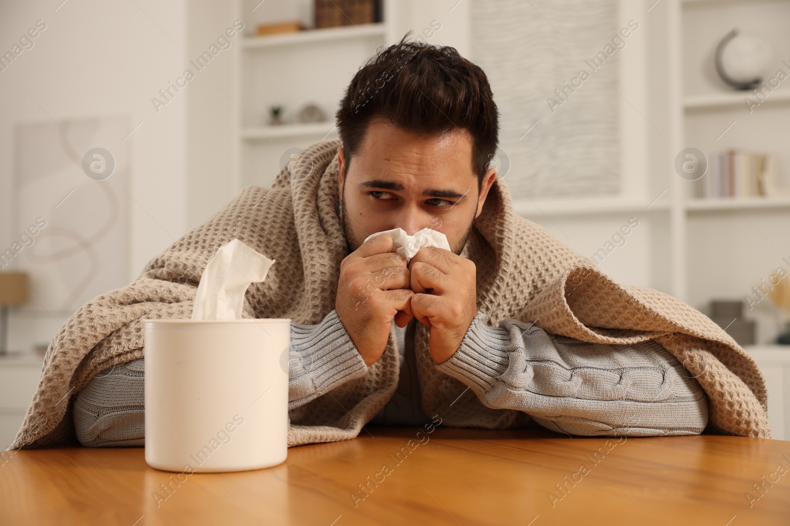 Photo of Sick man wrapped in blanket with tissue blowing nose at wooden table indoors. Cold symptoms