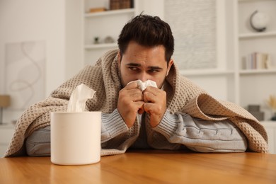 Photo of Sick man wrapped in blanket with tissue blowing nose at wooden table indoors. Cold symptoms