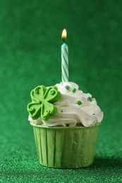 Photo of St. Patrick's day party. Tasty cupcake with clover leaf topper and burning candle on shiny green surface, closeup