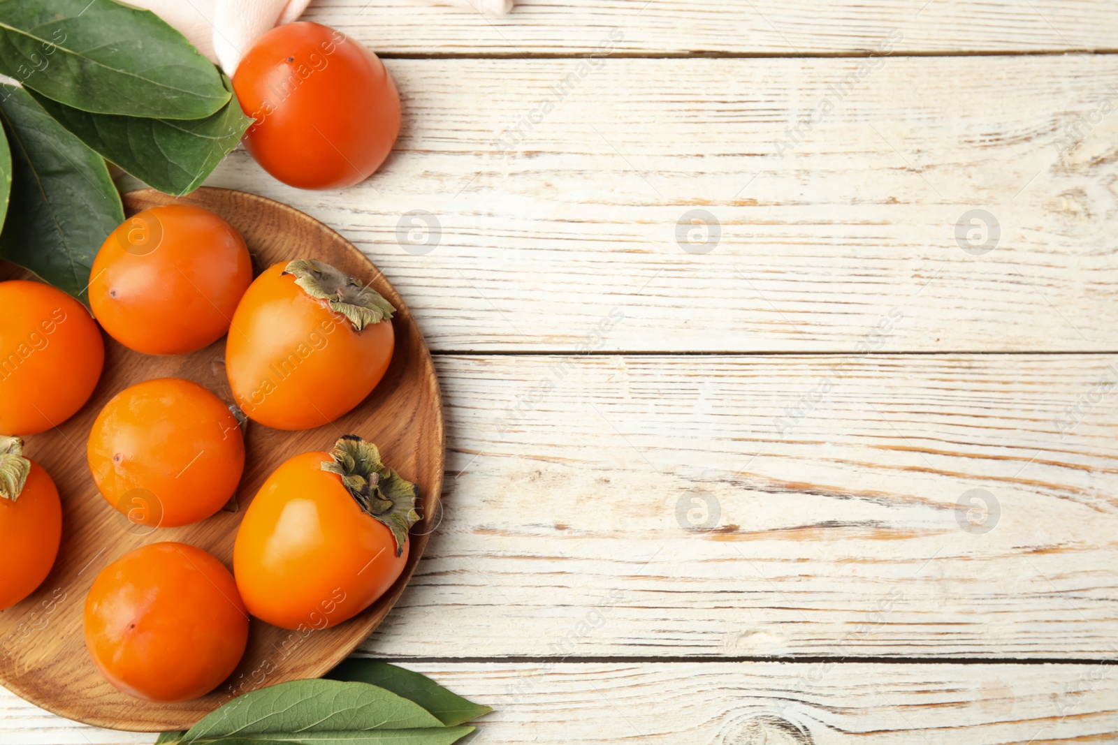 Photo of Delicious fresh persimmons and green leaves on white wooden table, flat lay. Space for text