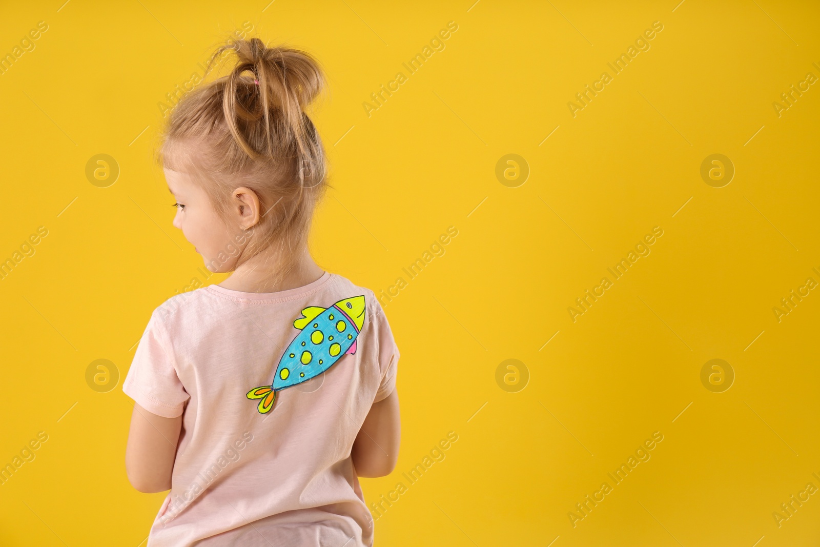 Photo of Little girl with paper fish on back against yellow background, space for text. April fool's day