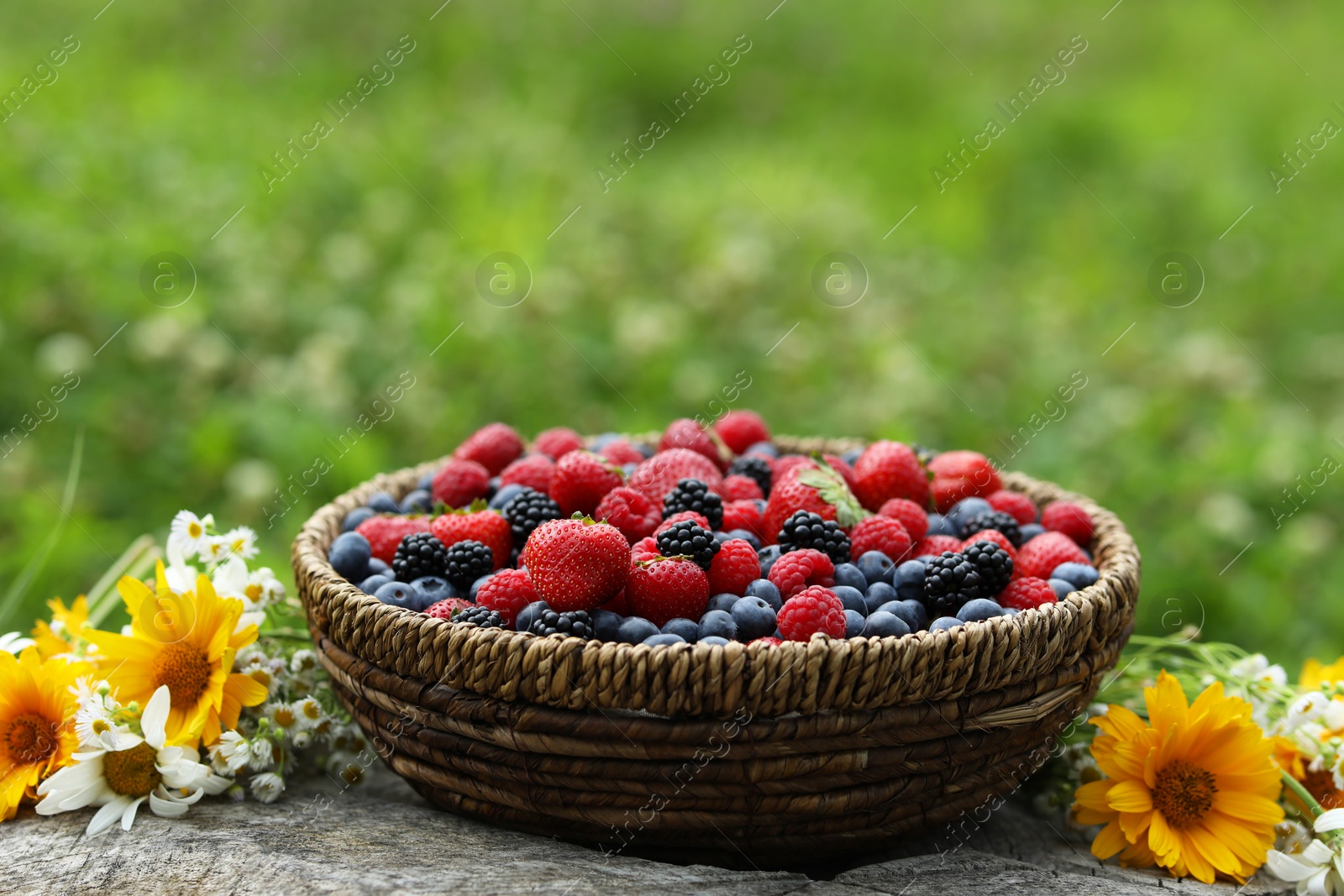 Photo of Wicker bowl with different fresh ripe berries and beautiful flowers on wooden surface outdoors