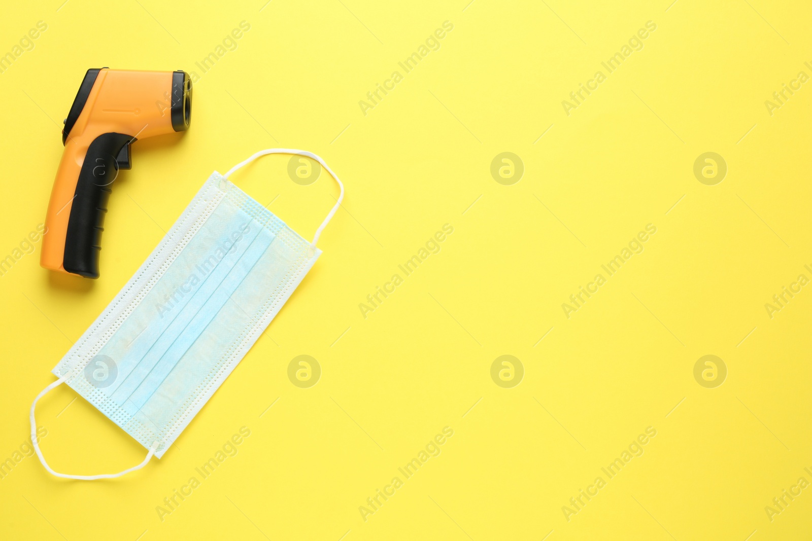 Photo of Infrared thermometer and medical mask on yellow background, flat lay with space for text. Checking temperature during Covid-19 pandemic
