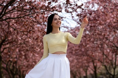 Photo of Pretty young woman wearing trendy clothes near beautiful blossoming trees outdoors. Stylish spring look