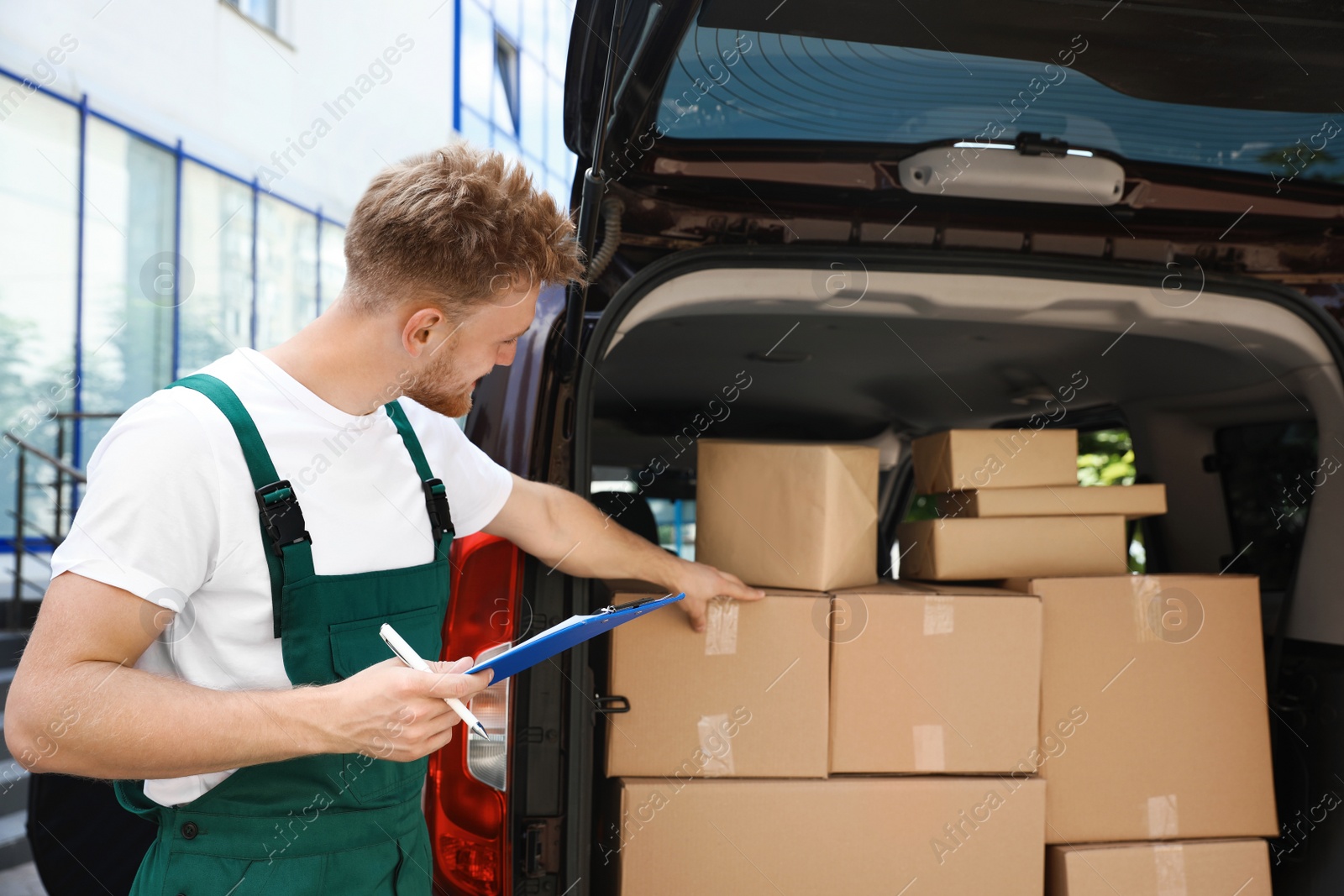 Photo of Young courier holding clipboard near delivery van with parcels outdoors