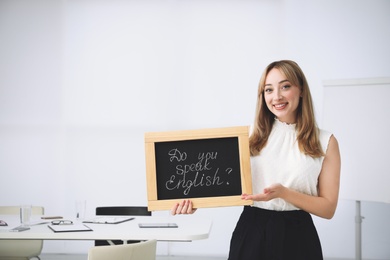 Photo of Young female teacher holding chalkboard with words DO YOU SPEAK ENGLISH? in classroom. Space for text