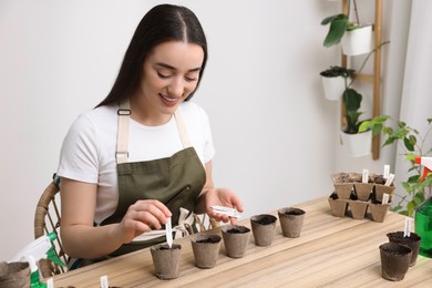 Photo of Woman inserting cards with names of vegetable seeds into peat pots at table indoors