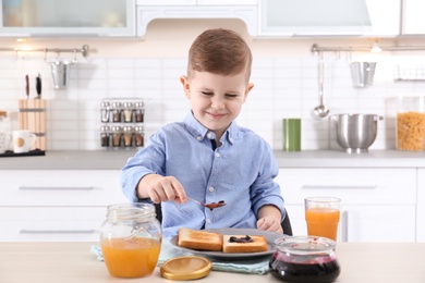 Photo of Cute little boy spreading sweet jam onto toast at table