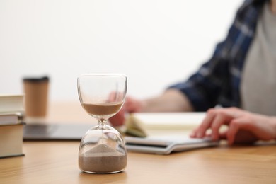 Photo of Hourglass with flowing sand on desk. Man taking notes indoors, selective focus