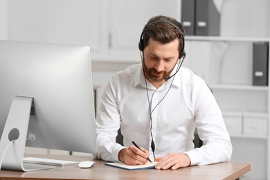 Photo of Hotline operator with headset writing something in notebook while working at wooden table