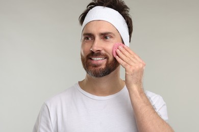 Photo of Man with headband washing his face using sponge on light grey background