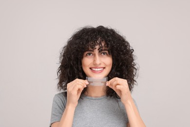 Young woman holding teeth whitening strips on light grey background