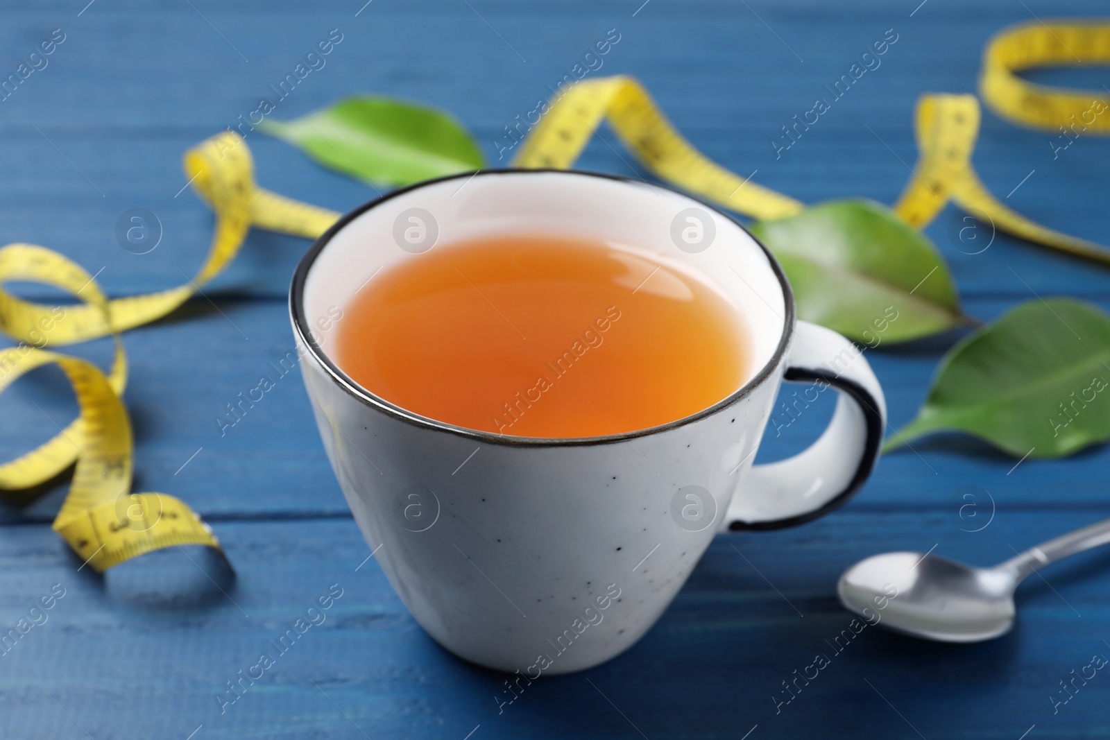 Photo of Cup of herbal diet tea, measuring tape, spoon and green leaves on blue wooden table, closeup. Weight loss concept