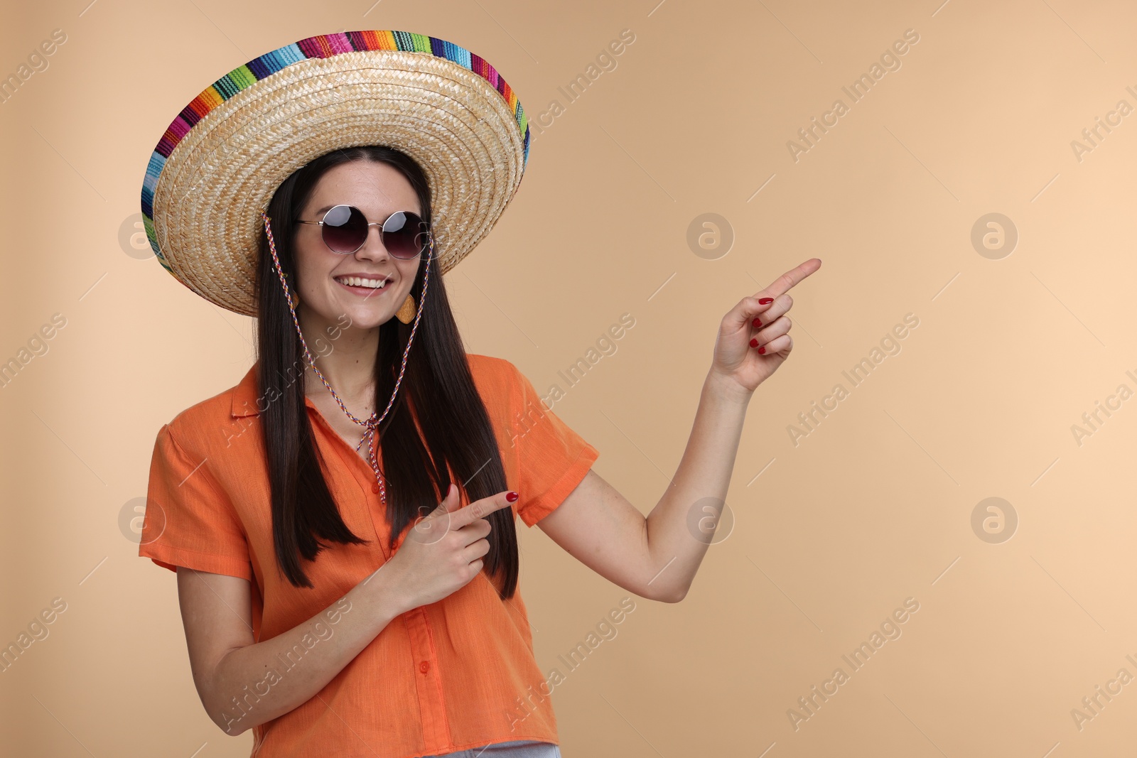 Photo of Young woman in Mexican sombrero hat and sunglasses pointing at something on beige background. Space for text