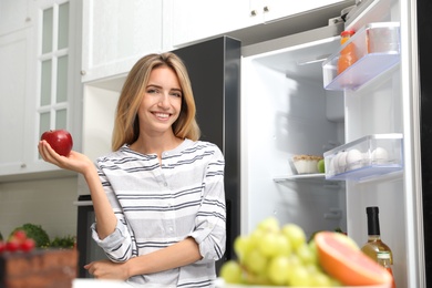 Photo of Concept of choice between healthy and junk food. Woman eating fresh apple near refrigerator in kitchen