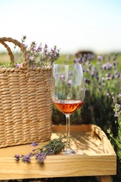 Photo of Glass of wine and wicker bag on wooden tray in lavender field