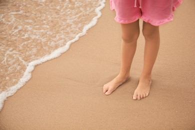 Photo of Little girl standing on sandy beach near sea, closeup. Space for text