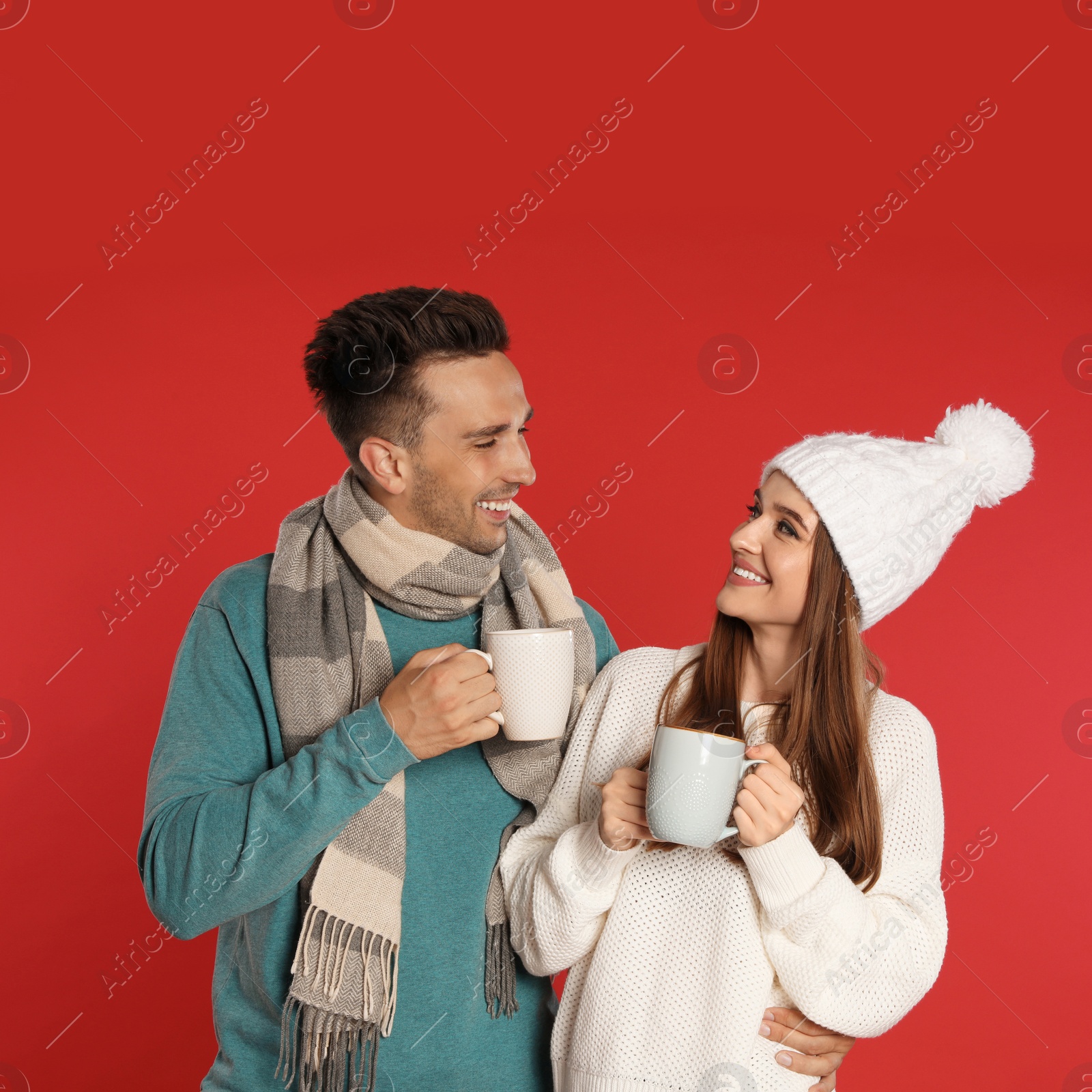 Photo of Happy young couple in warm clothes with cups on red background. Winter season