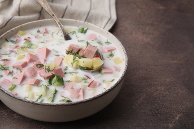 Delicious cold summer soup (okroshka) with boiled sausage and spoon in bowl on brown table. Space for text