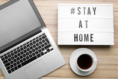 Photo of Laptop, cup of coffee and lightbox with hashtag STAY AT HOME on wooden background, flat lay. Message to promote self-isolation during COVID‑19 pandemic