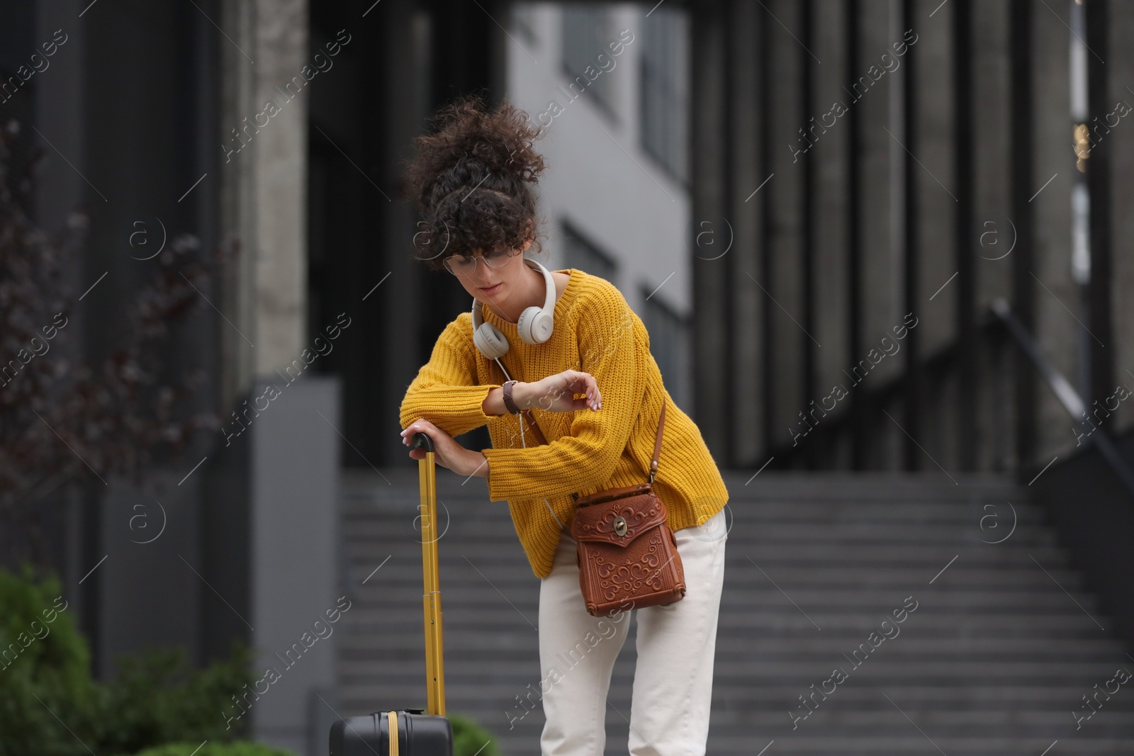 Photo of Being late. Worried woman with suitcase looking at watch near building outdoors