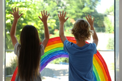 Photo of Little children near rainbow painting on window indoors. Stay at home concept