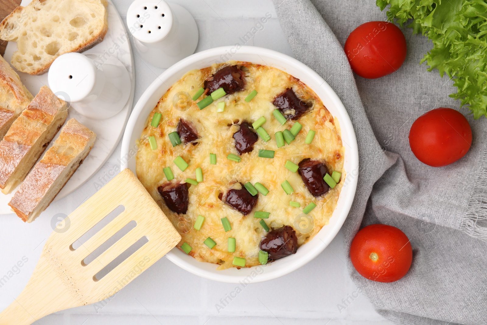 Photo of Tasty sausage casserole with green onions in baking dish served on white tiled table, flat lay