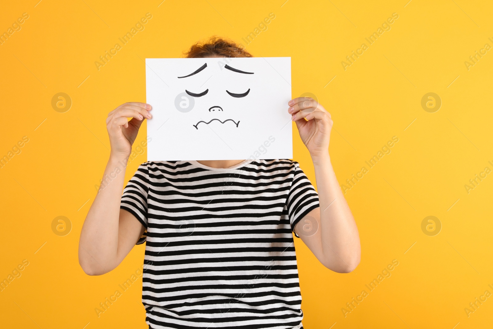 Photo of Woman hiding behind sheet of paper with sad face on yellow background