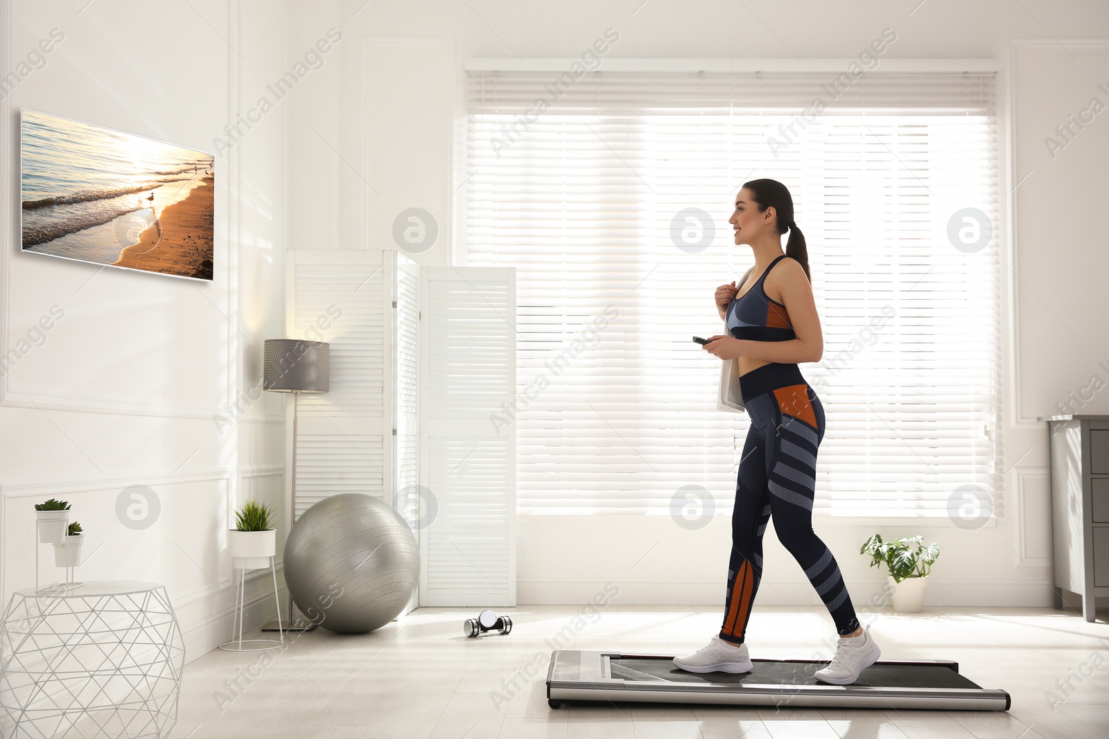 Image of Sporty woman with remote control training on walking treadmill and watching TV at home