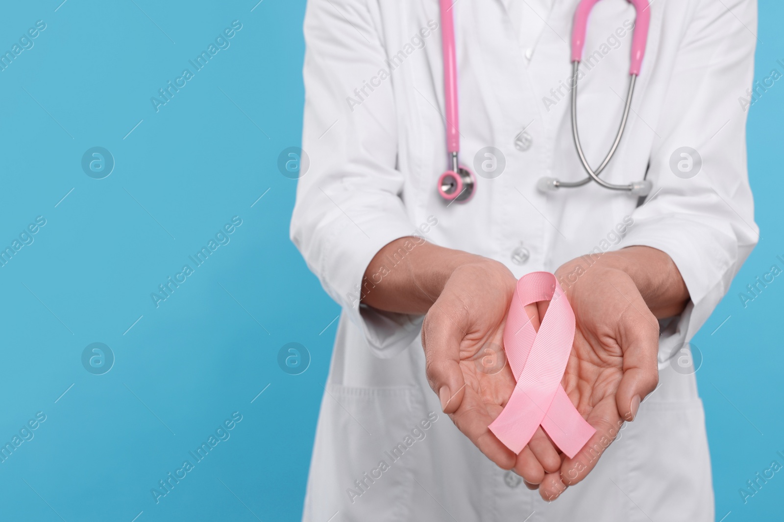 Photo of Doctor with stethoscope holding pink ribbon on light blue background, closeup and space for text. Breast cancer awareness