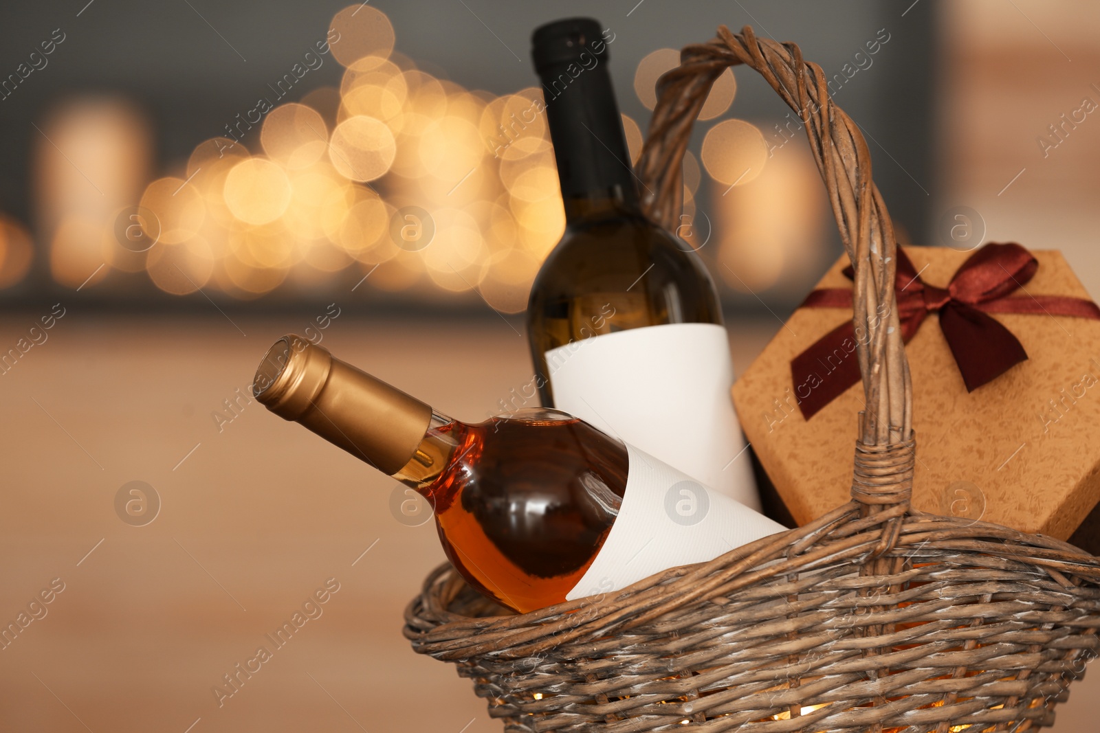 Photo of Wicker basket with bottles of wine and gift box against blurred background, closeup