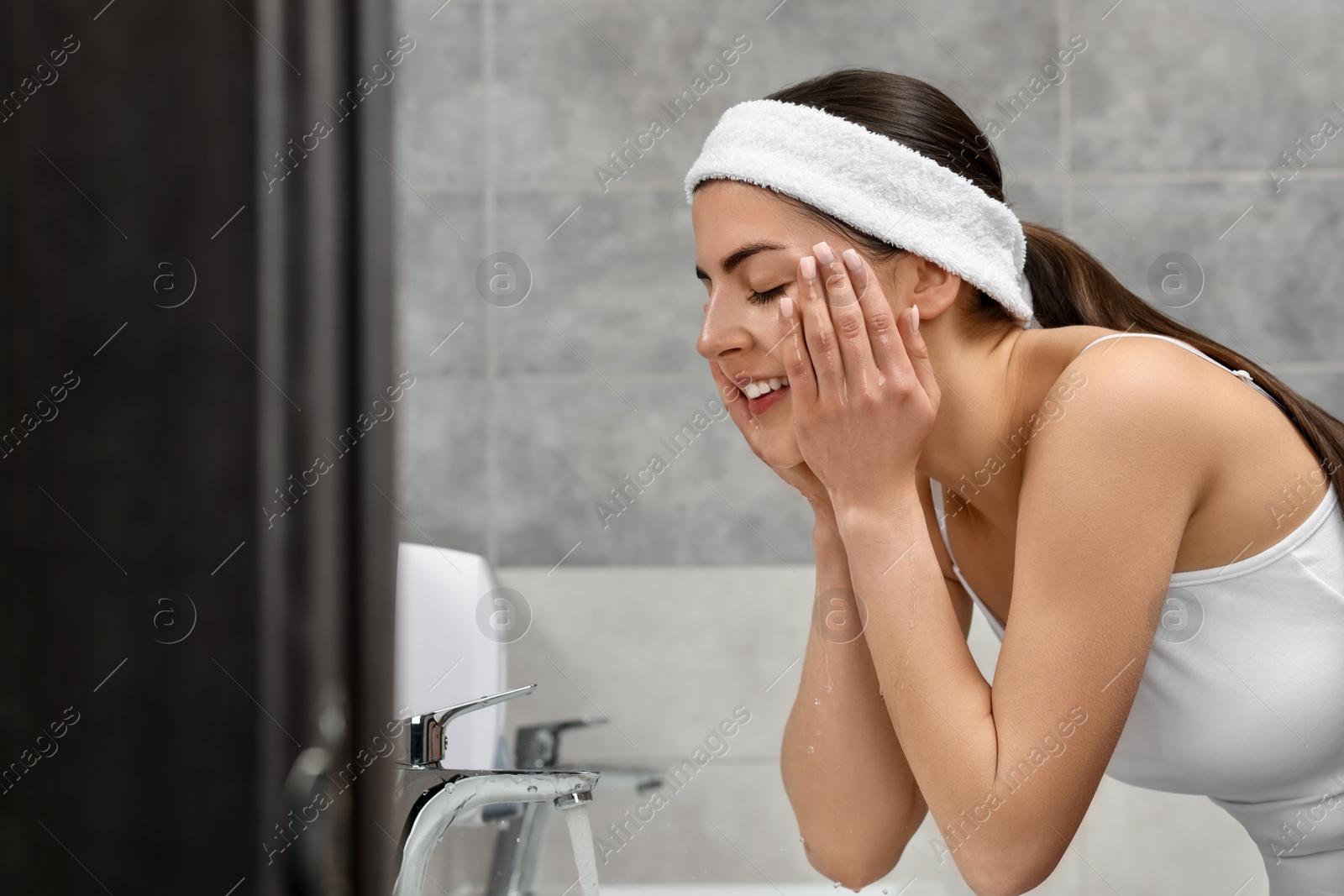 Photo of Young woman with headband washing her face in bathroom