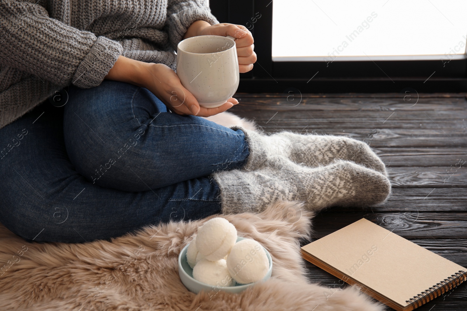 Photo of Woman in warm socks relaxing with cup of hot winter drink near window. Cozy season