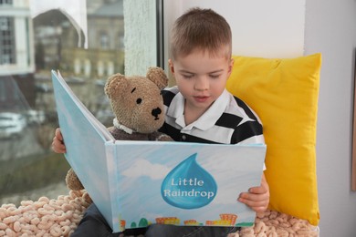 Photo of Cute little boy with toy bear reading book near window at home