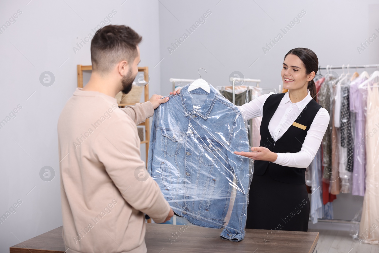 Photo of Dry-cleaning service. Happy worker giving denim jacket in plastic bag to client indoors