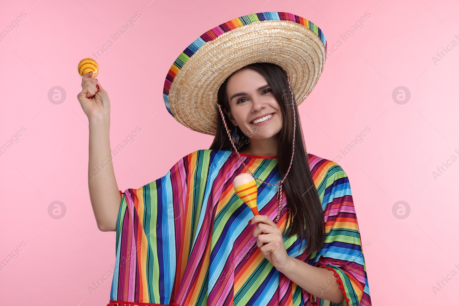 Photo of Young woman in Mexican sombrero hat and poncho dancing with maracas on pink background