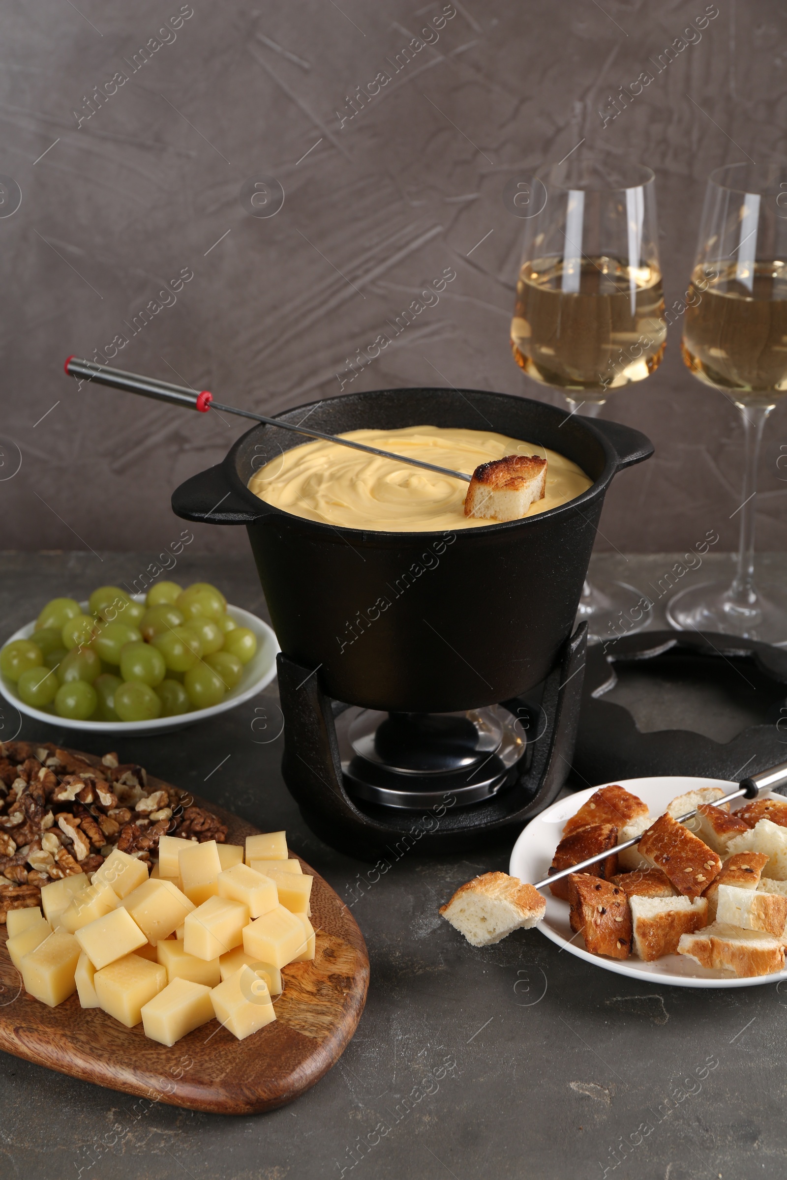 Photo of Fondue pot with tasty melted cheese, forks, wine and different snacks on grey table