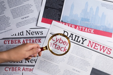 Photo of Woman using magnifying glass to read newspaper with headline CYBER ATTACK, top view