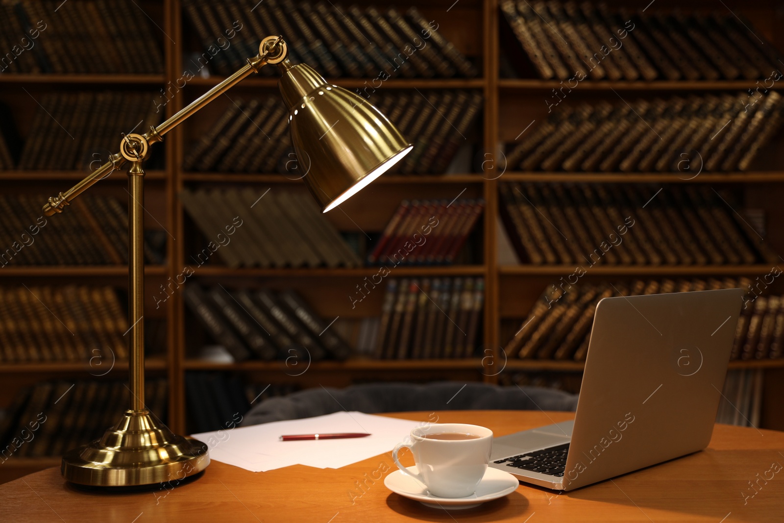 Photo of Lamp, cup of drink and laptop on wooden table near shelves with collection of vintage books in library