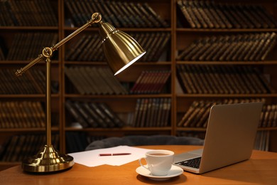 Lamp, cup of drink and laptop on wooden table near shelves with collection of vintage books in library