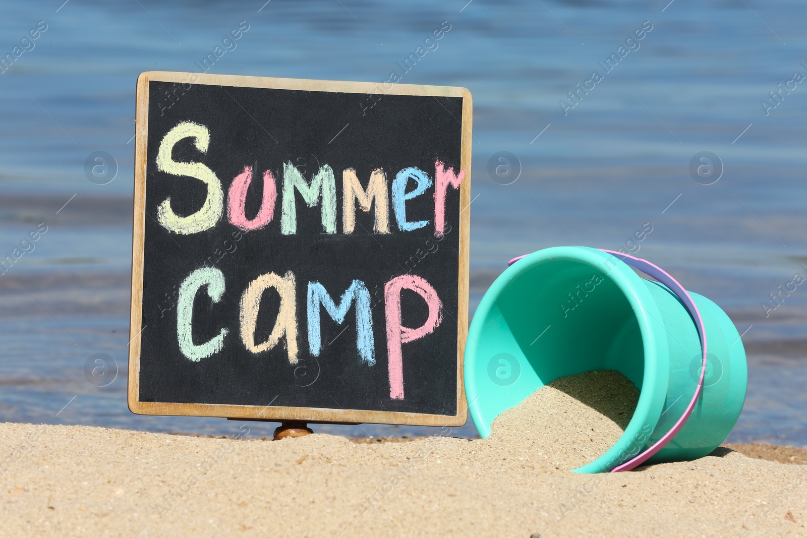 Photo of Little blackboard with text SUMMER CAMP and toy on sand near sea