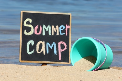 Photo of Little blackboard with text SUMMER CAMP and toy on sand near sea