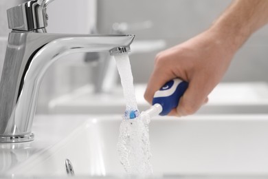 Photo of Man holding electric toothbrush under flowing water above sink in bathroom, closeup