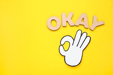 Word Okay made of wooden letters and paper cutout (OK hand gesture) on yellow background, flat lay. Space for text