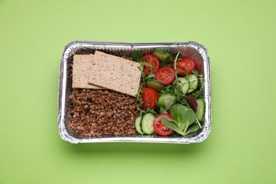 Photo of Container with buckwheat, fresh salad and crispbreads on light green background, top view