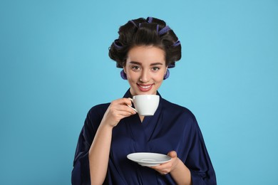 Photo of Happy young woman in silk bathrobe with hair curlers holding cup of drink on light blue background