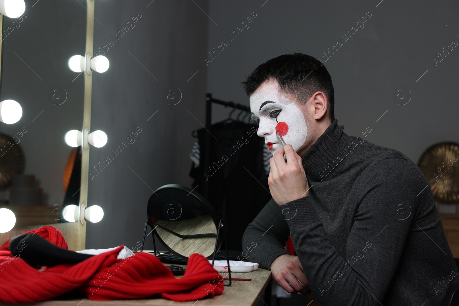 Photo of Young man applying mime makeup near mirror in dressing room