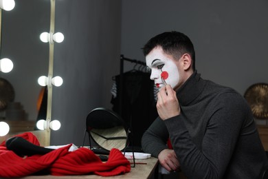 Photo of Young man applying mime makeup near mirror in dressing room