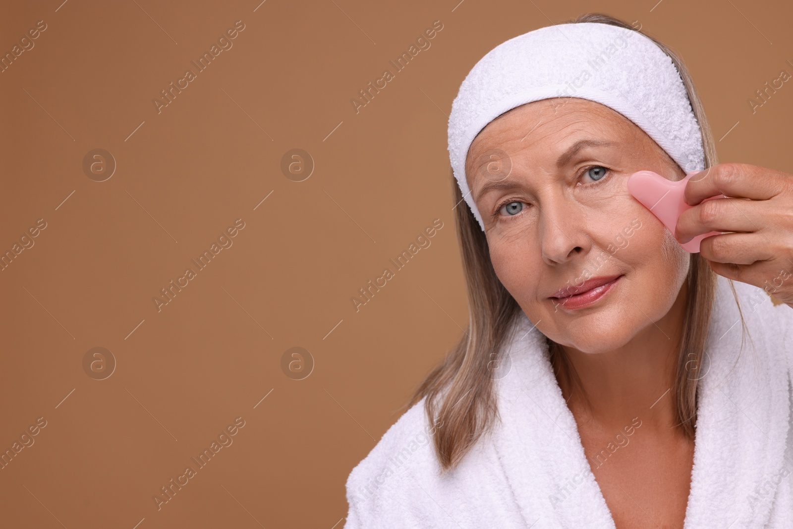 Photo of Woman massaging her face with rose quartz gua sha tool on brown background. Space for text